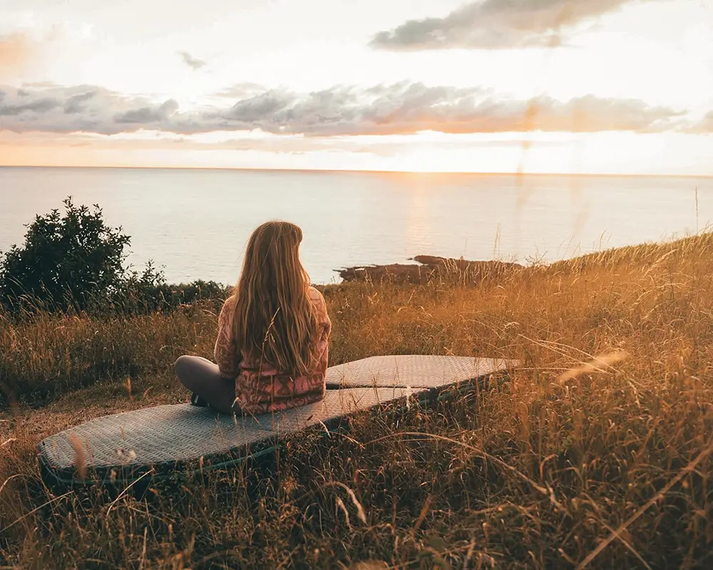 Frau sitzt auf einer Wiese mit einer Vanmade Matratze und blickt zum Meer, Abendstimmung